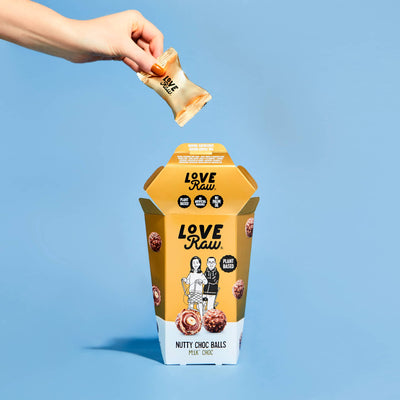 Dropping a gold-wrapped chocolate into LoveRaw Nutty Choc Balls container for vegan indulgence