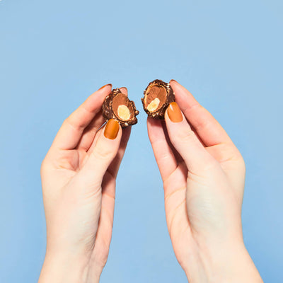 Hands holding broken LoveRaw Nutty Choc Balls showing hazelnut center, vegan chocolate indulgence